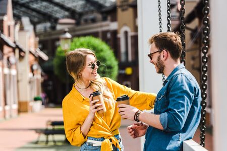cheerful woman in sunglasses looking at handsome man and holding paper cup