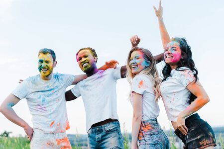 happy man with hand on hip near multicultural friends with holi paints on faces