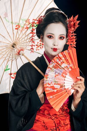 smiling beautiful geisha in black kimono with red flowers in hair holding traditional asian umbrella and hand fan isolated on blackの写真素材