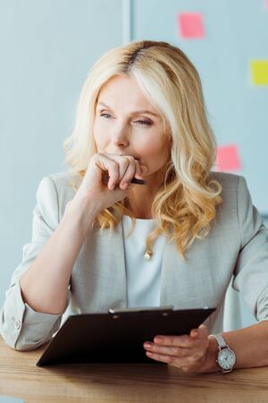 thoughtful blonde woman covering face while holding pen and clipboardの写真素材