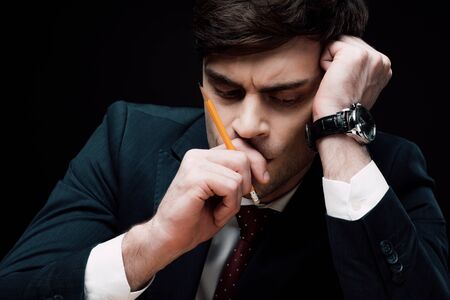 upset, thoughtful businessman holding hand with pencil near face isolated on blackの写真素材