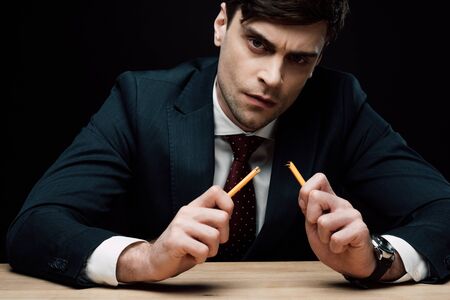 serious businessman holding broken pencil and looking at camera isolated on blackの写真素材