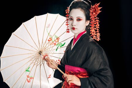 geisha in black kimono with red flowers in hair holding traditional asian umbrella isolated on blackの写真素材
