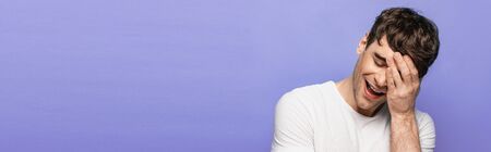 panoramic shot of sheerful man smiling with closed eyes while holding hand on face isolated on blueの写真素材