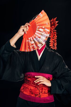beautiful geisha in black kimono with red flowers in hair holding traditional hand fan in front of face isolated on blackの写真素材