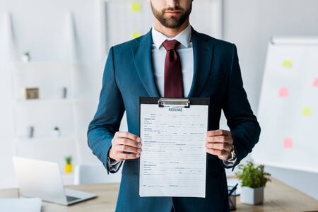 cropped view of bearded recruiter in suit standing and holding clipboard with resume letters