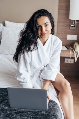 charming brunette young woman in white bathroom using laptop in bedroomの写真素材
