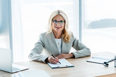 cheerful blonde woman in glasses looking at clipboard near laptopの写真素材