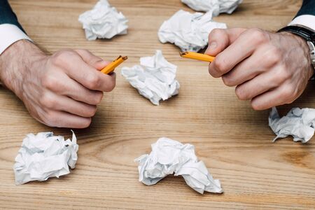 cropped view of businessman holding broken pencil while sitting at wooden desk with crumpled pape