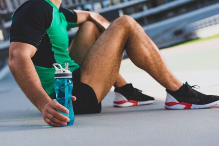 cropped view of sportsman sitting on running track and holding bottle with waterの写真素材
