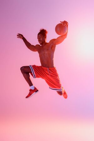handsome, muscular african american sportsman in red shorts jumping while playing basketball on pink and purple gradient backgroundの写真素材