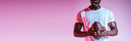 partial view of african american sportsman screaming while holding rugby ball on purple background with gradient, panoramic shotの写真素材