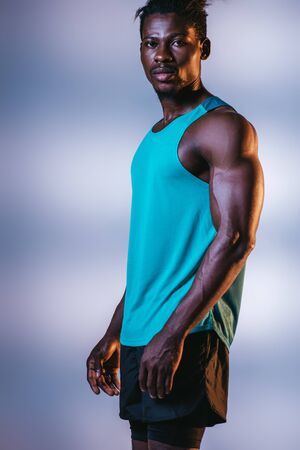 handsome, athletic african american sportsman looking at camera on grey and blue background with lightingの写真素材