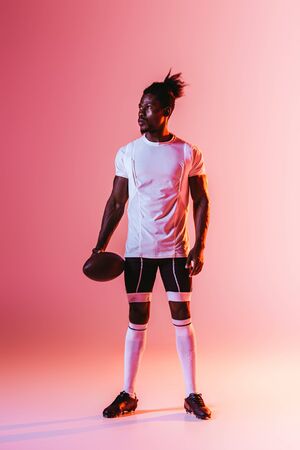 handsome african american sportsman looking away while holding rugby ball on pink background with gradient and lightingの写真素材