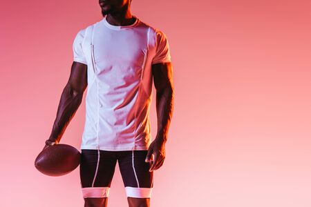 cropped view of african american sportsman holding rugby ball on pink background with gradient and lightingの写真素材