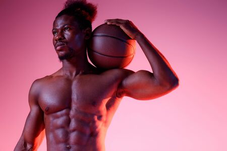 serious african american sportsman with muscular torso holding ball and looking away on pink and purple gradient backgroundの写真素材