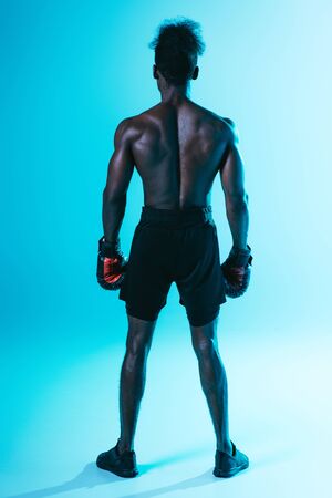 back view of shirtless, muscular african american sportsman in boxing gloves on blue backgroundの写真素材
