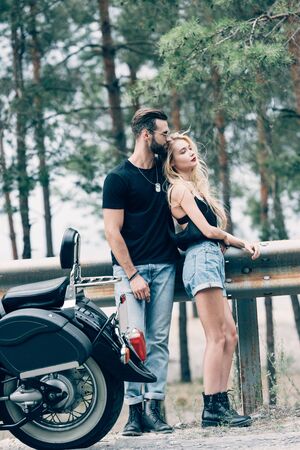 young couple of bikers embracing near black motorcycle on road near green forestの写真素材