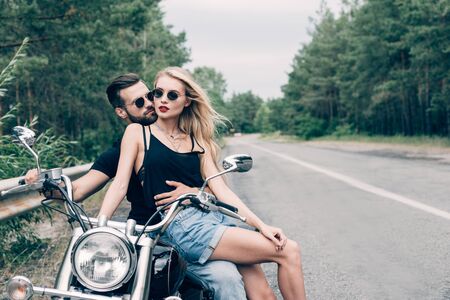 young couple of bikers closely sitting on black motorcycle on road near green forestの写真素材