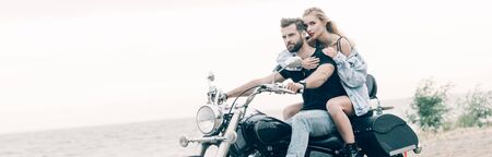 young loving couple of bikers sitting on black motorcycle at beach near river, panoramic shotの写真素材
