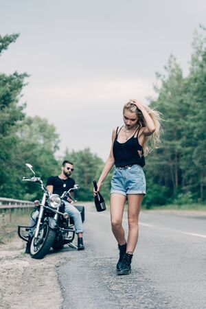 selective focus of young woman walking on road with bottle of alcohol near boyfriend on black motorcycleの写真素材