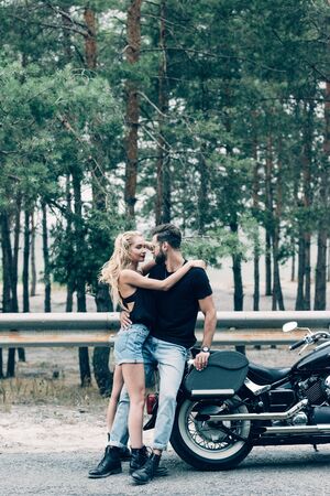 sexy young couple of bikers hugging near black motorcycle and green forestの写真素材