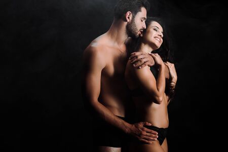 shirtless man kissing happy young woman standing on black with smoke