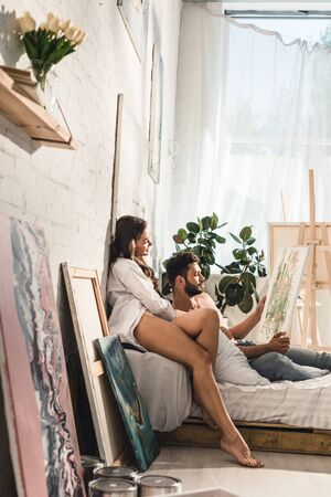 side view of sexy young couple looking at painting while man lying in bed and girl sitting next to guyの写真素材