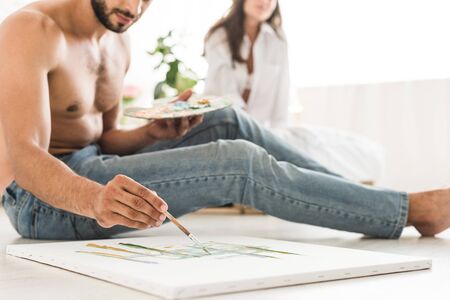 cropped view of man drawing with brush while sitting on floor and girl sitting in bedの写真素材