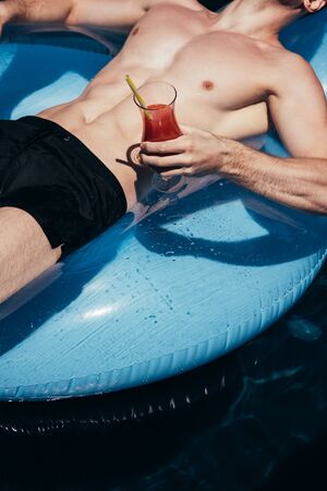 partial view of young man relaxing on swim ring in swimming pool with glass of refreshing drink