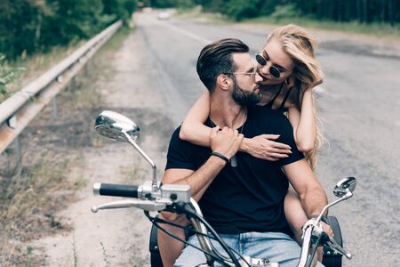 young couple of bikers hugging and looking at each other on black motorcycle on road near green forestの写真素材