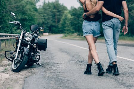 cropped view of young couple of bikers walking along road and embracing near black motorcycleの写真素材