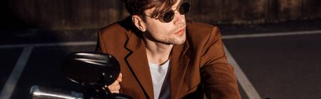 panoramic shot of handsome man in sunglasses sitting on motorcycle and looking awayの写真素材