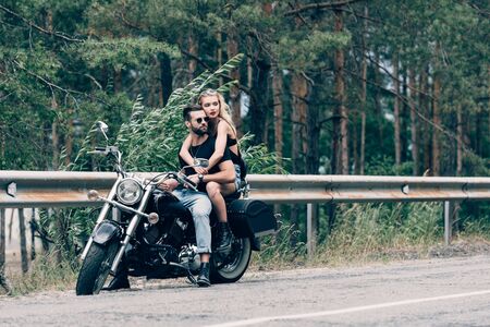 young couple of bikers hugging while sitting on black motorcycle on road near green forestの写真素材