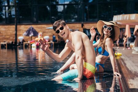 cheerful young couple in sunglasses waving hands at camera while relaxing on poolside
