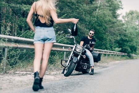 back view of young woman walking on road with bottle of alcohol near boyfriend on black motorcycle on roadの写真素材