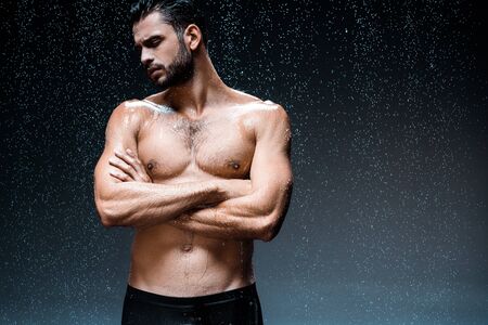 handsome wet man standing with crossed arms under raindrops on blackの写真素材