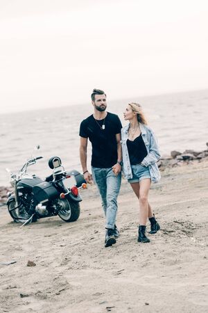 young couple of bikers in love walking and holding hands near black motorcycle at sandy beachの写真素材