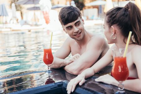 cheerful young couple looking at each other while resting at poolside with glasses of refreshing drinkの写真素材