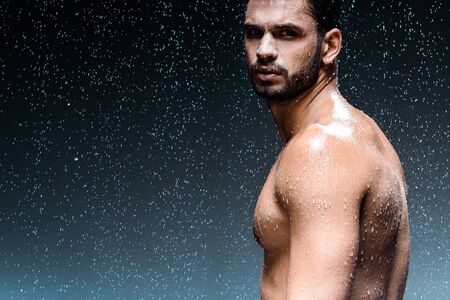 wet man looking at camera while standing under raindrops on blackの写真素材