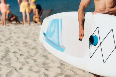 cropped view of young man holding surfing board while standing on beachの写真素材