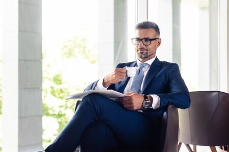 handsome businessman in suit and glasses holding cup and newspaperの写真素材