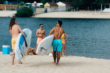 back view of young multicultural men holding surfing boards on beachの写真素材