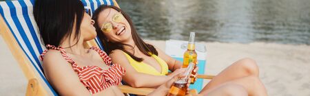 panoramic shot of cheerful, beautiful multicultural girls sitting in chaise lounges with bottles of...