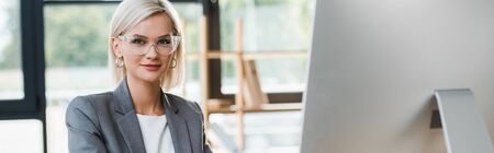 panoramic shot of cheerful blonde woman in glasses looking at camera in officeの写真素材