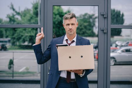 handsome man in suit showing middle finger while holding box near buildingの写真素材