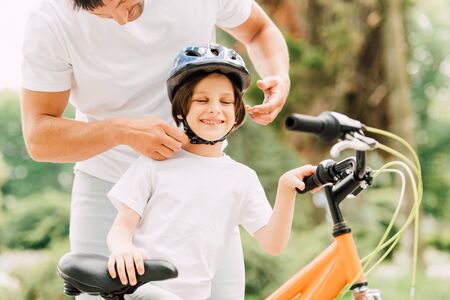 cropped view of father putting helmet on son while boy smiling and standing near bicycleの写真素材