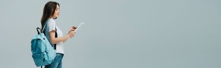 side view of pretty smiling girl with blue backpack using digital tablet isolated on grey, panoramic shotの写真素材