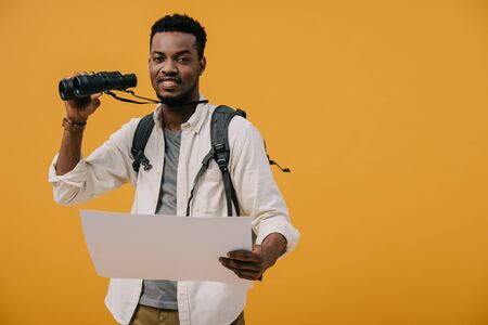 happy african american man holding binoculars and blank paper isolated on yellow