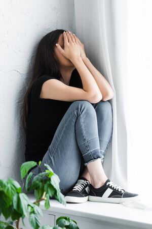 depressed young woman crying while sitting on window sill and covering face with handsの写真素材